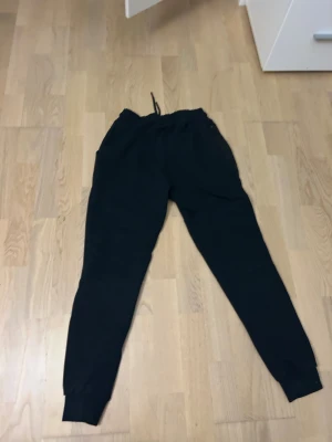 Svarta mjukisbyxor från Nike, strl M - Svarta mjukisbyxor från Nike med diskret logga på benet. Byxorna har snörning i midjan, ribbade muddar vid bensluten och två sidofickor. Perfekta för chill eller träning, tillverkade i mjukt bomullsmaterial för maximal komfort.