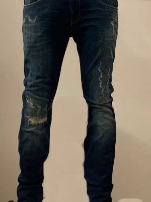 Replay Anbass jeans blå, waist 28/32 - Snygga Replay Anbass jeans i mörkblå tvätt med slitningar och distressed detaljer på framsidan. Modellen har smal passform, klassiska fem fickor och kontrastsömmar. Materialet är mjukt jeans-tyg med stretch för extra komfort.