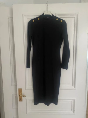 Svart ribbad långklänning Degaje, ny utan prislapp - Svart ribbad långklänning från Degaje med lång ärm och höga axlar dekorerade med fyra guldknappar. Klänningen har en figurnära passform och hög hals, perfekt för en stilren look.