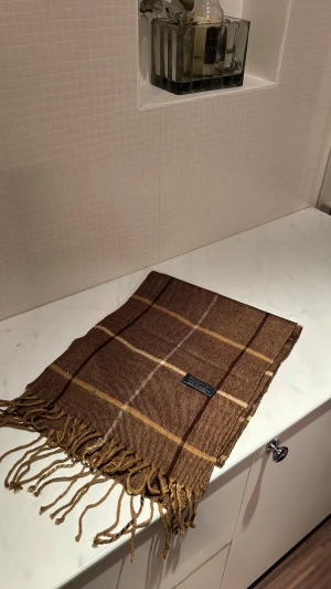 Brun rutig ullväst från Burberrys - Snygg brun väst från Burberrys i 100% lammull, med klassiskt rutigt mönster i beige och mörkbruna toner. Västen har fransar längs nederkanten och en mjuk, varm känsla som passar perfekt för kyliga dagar. Engelsk design med exklusiv känsla.