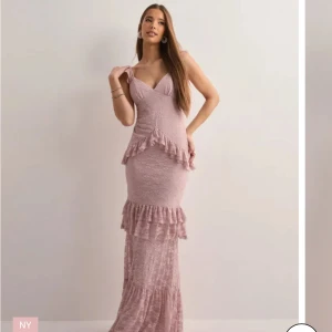 Ljusrosa spetsklänning med volanger - Elegant ljusrosa långklänning i spets med volangdetaljer och öppen rygg. Klänningen har smala axelband och en v-ringad urringning. Perfekt för speciella tillfällen.