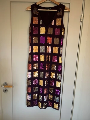 Glittrig klänning från Karen Millen - Snygg långklänning från Karen Millen med glittriga paljettrutor i guld, lila, rosa och silver på mörklila botten. Klänningen är ärmlös, har v-ringning och en figurnära passform. Perfekt för dig som vill sticka ut med färg och glitter.