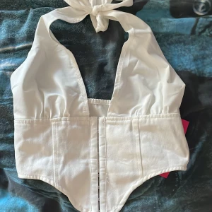 Vit korsettopp med knytband - Säljer en helt ny endast testad vit korsettopp från SHEIN i storlek xxs med knytband i nacken och markerad midja. Toppen har en halterneck-modell och stängning framtill. Den kom me en matchade kjol men tyväär var korsetten för liten för mig.