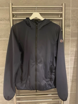 Mörkblå vindjacka från Colmar - Snygg mörkblå vindjacka från Colmar med luva och hel dragkedja framtill. Jackan har två sidofickor med dragkedja och en rund Colmar-logga på ärmen. Perfekt för dig som gillar stilrena och sportiga plagg. Materialet är lätt och syntetiskt. Har ni några frågor tveka inte av att höra av er!