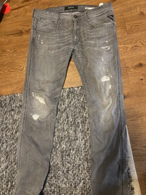 Replay Anbass grå slim jeans 33 - Replay Anbass jeans i grå tvätt med slim passform. Snygga slitningar och trasiga detaljer på benen ger en cool, avslappnad vibe. Klassisk femficksmodell med svarta sömmar på bakfickan. Perfekt för dig som gillar streetstyle.