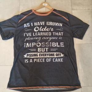 Mörkgrå t-shirt med tryck och kontrastkrage - Säljer en mörkgrå t-shirt i polyester med ett stort, humoristiskt vitt tryck på framsidan. T-shirten har korta ärmar och en kontrastfärgad krage i brunorange. Perfekt för dig som gillar att sticka ut med roliga budskap.