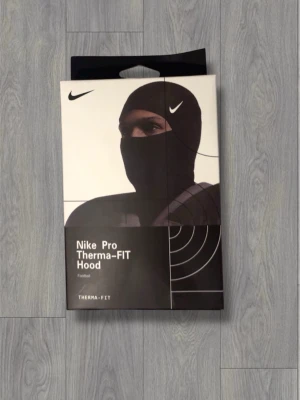 Nike Pro Therma-FIT svart huva - Nike Pro Therma-FIT Hood i svart, perfekt för fotboll och träning. Huvan är tillverkad i mjuk och stretchig polyester med platta sömmar för extra komfort. Snyggt Nike Pro-märke framtill och vit Swoosh på sidan. Passar både under hjälm och som standalone.