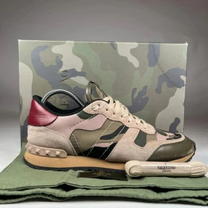 Valentino Rockrunner sneakers camouflage - Snygga Valentino Rockrunner sneakers med beige, olivgrön och rosa camouflagemönster, röda detaljer vid hälen och ikoniska nitar. Skorna har snörning, platt sula och är tillverkade i mocka och skinn med textilinslag. Perfekt för dig som vill sticka ut med exklusiv streetstyle.