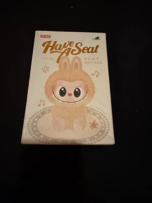 Labubu Have A Seat Baba från Pop Mart Äkta - Säljer en beige pälsväst inspirerad av Labubu från Pop Mart. Västen har en fluffig look med rundad form och söta kaninöron på huvan. Perfekt för dig som gillar gulliga och unika plagg med mjuk pälsfeeling. Färgen är ljusbeige och designen är riktigt charmig.