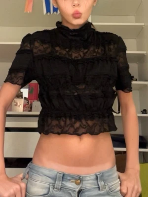 Svart  croppad blus med volanger - Säljer en svart och croppad blus med korta ärmar och hög hals. Blusen har volanger och är gjord i ett tunt spetsmaterial. Säljer då den inte andvänds, har andvänt den ungefär 2-3 gånger. Är 165 och den sitter bra🥰 vet tyvärr inte märke/storlek då det inte står men skulle gissa att storlek är XS.