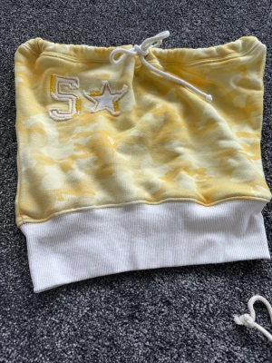 Gul och vit camo tubtopp med snörning - Säljer en tubtopp i ljusgul och vit camo med bred vit ribbad kant nedtill och snörning upptill. Broderad siffra 5 och stjärna framtill i vitt och gult. Perfekt för dig som vill sticka ut med en cool och sportig vibe.