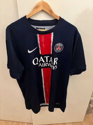 PSG Nike fotbollströja marinblå - Snygg PSG fotbollströja från Nike i marinblått med röd och vit randig detalj på framsidan. Tröjan har klubbmärke på bröstet och Qatar Airways tryck. Perfekt för dig som älskar fotboll och vill visa ditt stöd för Paris Saint-Germain.
