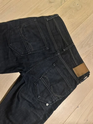 Mörkblå jeans från Jack & Jones (slim fit/GLENN) -  Snygga mörkblå jeans från Jack & Jones Modell slim fit/GLENN med klassisk femficksdesign och kontrastsömmar. Modellen har rak passform och normal midja, tillverkad i slitstarkt denim. Perfekt för dig som gillar stilrena och tidlösa jeans. W29 L32