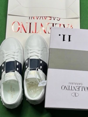 Valentino Garavani vita sneakers - KAN DUSKITERAS OM PRIS! Snygga vita sneakers från Valentino Garavani med bred mörkblå rem över ovansidan och coola nitar längs sulan. Skorna har rund tå, platt sula och klassisk snörning. Märkeslogga i guld på plösen och insidan. Perfekta för dig som gillar exklusiv streetstyle.