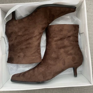 Bruna mocka boots med klack - Snygga bruna stövletter i mocka med spetsig tå och låg klack. Dem är helt nya och har aldrig använts. Skorna har en elegant siluett och passar perfekt till både jeans och kjol. Materialet är mjukt och ger en lyxig känsla. Perfekta för dig som vill ha en stilren och trendig look.