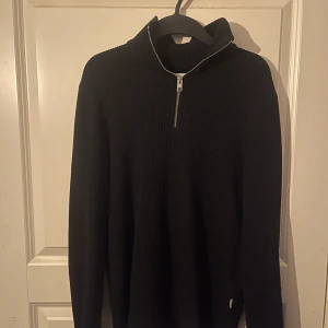 Svart stickad half zip från Jack & Jones - Svart stickad tröja från Jack & Jones med ribbad struktur och half zip-dragkedja vid halsen. Tröjan har lång ärm och en hög krage som ger en clean och stilren look. Perfekt för dig som gillar enkel och snygg design.