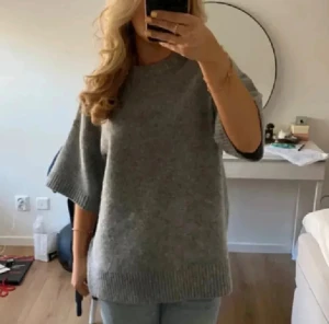 Grå stickad tröja med kort ärm - Säljer en grå stickad tröja med korta, vida ärmar och rund halsringning. Tröjan har ribbade muddar vid ärmslut och nederkant, vilket ger en snygg och enkel look. Perfekt att styla med jeans eller kostymbyxor.