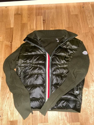 Moncler grön pufferjacka med stickade ärmar - Snygg pufferjacka från Moncler i mörkgrönt med stickade ärmar och hög krage. Jackan har en quiltad front i glansigt material, dragkedja med rödvit detalj och Moncler-logga på ärmen. Perfekt för dig som vill ha en sportig och trendig look.
