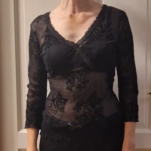 Svart blommig meshblus från KappAhl - Säljer en transparent svart meshblus från KappAhl med blommigt sammetsmönster. Blusen har v-ringning med spetskant och trekvartsärm.