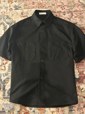 Svart kortärmad skjorta från Zara - Svart skjorta från Zara med korta ärmar, klassisk krage och två stora bröstfickor. Skjortan har knappar framtill och en clean, minimalistisk look som passar perfekt till jeans eller shorts.