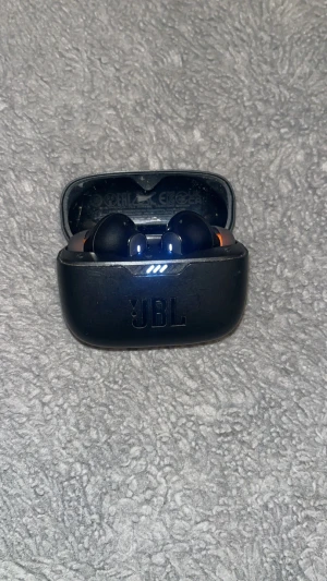 JBL True Wireless Earbuds - JBL trådlösa hörlurar med laddningsetui. Kompakt design, perfekt för musik och samtal på språng. Laddningsindikator synlig, viss kosmetisk slitage på etuiet men fullt fungerande. Levererar JBL:s signaturljud och stabil anslutning.