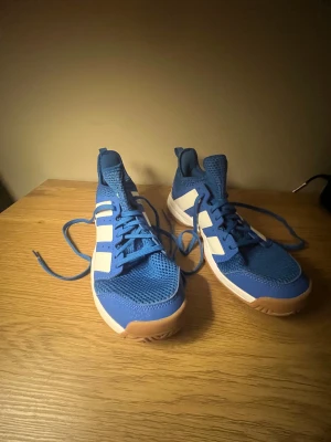 Blå Adidas sneakers Inomhusskor  - Säljer ett par sportiga Adidas sneakers i blått med vita ränder och detaljer. Ovandelen är i ventilerande mesh och syntet, med snörning och platt gummisula i beige. Perfekt för träning eller häng, med coola nion gröna detaljer på hälen och klassisk Adidas-design. Passar bra för inomhus sport/träning.