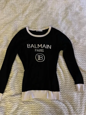 Svart stickad tröja från Balmain - Svart stickad tröja från Balmain Paris med vit logotyp framtill. Tröjan har vita ribbade detaljer vid halsringning, ärmslut och nederkant. Slim passform och långärmad, perfekt för dig som gillar exklusiva märken och stilren design.