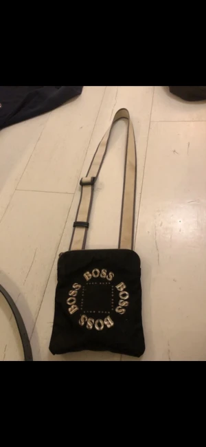 Svart axelväska från Hugo Boss - Svart axelväska äkta köpt från Hugo Boss med justerbar beige axelrem. Lite smutsigt band men går att tvätta Väskan har tydlig BOSS-logga i vitt tryck på framsidan och en fyrkantig form. Perfekt för att bära det viktigaste och ge en cool touch till din outfit.