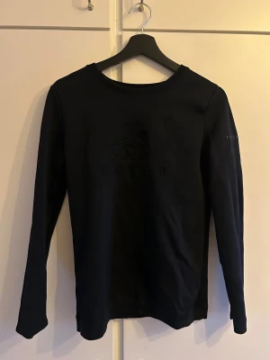 Svart långärmad tröja från Fager - Svart långärmad tröja från Fager med diskret logotyp i relief på bröstet och tryck på ärmen. Tröjan har en klassisk rund hals och är gjord i mjuk bomull som känns skön mot huden. Perfekt för dig som gillar stilrena och enkla plagg.