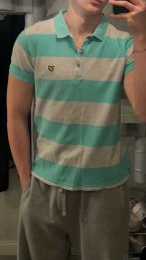 Randig polotopp från Lyle & Scott - Säljer en fräsch polotopp från Lyle & Scott med breda turkosa och ljusgrå ränder. Toppen har korta ärmar, klassisk krage och tre knappar framtill. På bröstet sitter den ikoniska gula fågeln broderad. Perfekt för dig som gillar sportig och chill stil.