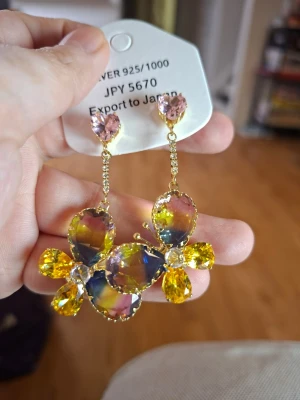 Färgglad fjärilsväst med glittriga stenar örhängen  - Säljer en unik örhängen  med stora glittriga fjärilar i gult, rosa och blått. Örhängen har en cool design med stenar i olika färger och former, och detaljer i guld. Perfekt för dig som vill sticka ut och älskar färg och bling.