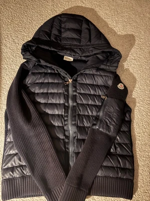 Mörkblå stickad cardigan jacka Moncler - Säljer en mörkblå stickad cardigan jacka från Moncler i storlek M. Jackan har Moncler-logga på ärmen, ribbad stickning och detaljer i syntetmaterial vid muddar och insida. Perfekt för dig som gillar exklusiva märken och snygg design.