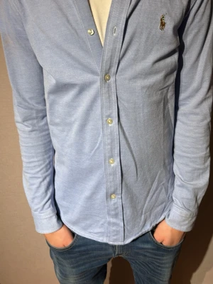 Ljusblå skjorta från Ralph Lauren - Säljer en stilren ljusblå skjorta från Ralph Lauren med klassisk knappstängning och broderad logga på bröstet. Skjortan är i mjuk bomull och har lång ärm samt en clean, tidlös look som funkar till jeans eller chinos.