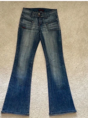 Blå bootcut jeans från DCH Jeans - Säljer ett par blå bootcut jeans från DCH Jeans med coola detaljer som dubbla knappar i midjan, framfickor med lock och bakfickor med dragkedja. Jeansen har en snygg tvätt med ljusa slitningar och vita vertikala ränder i tyget.