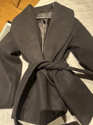 Svart kappa från Vero Moda med bälte - Säljer en svart kappa från Vero Moda med bred krage och knytbälte i midjan. Jackan har en klassisk och stilren look, perfekt för kyligare dagar. Materialet känns mjukt och har en ullig finish. Passformen är normal och ärmarna är långa.