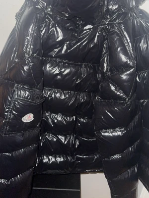 Svart Moncler Maya pufferjacka ( pris går att diskutera ) - Säljer en svart, glansig pufferjacka från Moncler med klassisk logga på ärmfickan. Jackan har quiltad design, dragkedja och stora fickor. Perfekt för kalla dagar och riktigt snygg till streetwear-stil.