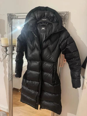 Svart lång pufferjacka Mountain Works XS - Säljer en svart lång pufferjacka från Mountain Works i storlek XS. Jackan har huva, dragkedja framtill och snygga quiltade detaljer. Perfekt för kalla dagar med sin fluffiga och varma känsla. Snygg passform med dragsko i midjan och praktiska fickor. Helt ny 