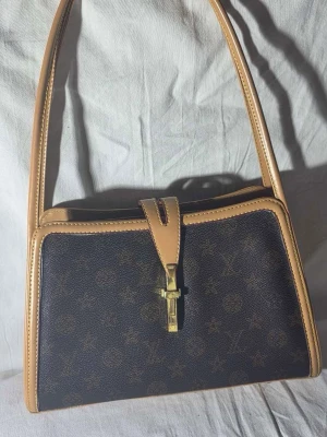 Louis Vuitton axelväska monogram - Säljer en snygg axelväska från Louis Vuitton med klassiskt monogrammönster i mörkbrunt och beige. Väskan har guldfärgad metallstängning och detaljer i skinn. Perfekt storlek för det viktigaste och har en stilren, rektangulär form med långa axelremmar. Köpt på vinted för 5000kr 