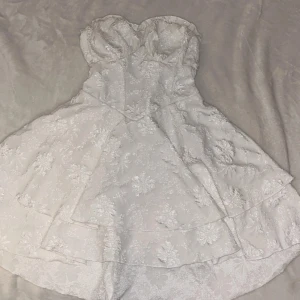 Vit axelbandslös klänning med blommigt mönster - Charmig vit axelbandslös klänning som formar midjan med ett vackert blommigt mönster. Klänningen har en volangdetalj Den har en kort längd och en elegant passform som framhäver figuren. Bust:74-100 cm, Waist Size:67-96 cm, Length:64 cm