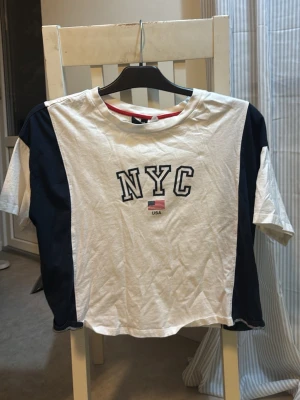 Vit och marinblå NYC t-shirt - Säljer en snygg t-shirt med NYC-tryck och liten USA-flagga på bröstet. T-shirten är vit med marinblå paneler på sidorna och ärmarna. Lite croppad stil