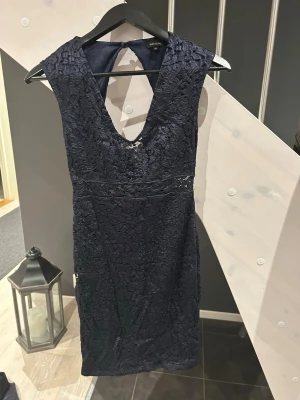 Mörkblå spetsklänning  - Snygg mörkblå kortklänning från River Island med v-ringning och öppen rygg. Klänningen är ärmlös och har vackra spetsdetaljer över hela tyget samt en dragkedja bak. Perfekt för dig som vill sticka ut med en elegant look.