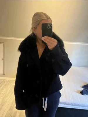 Svart teddyjacka med fuskpäls från Meotine - Säljer en svart teddyjacka från Meotine med stor fluffig fuskpäls-krage. Jackan har en boxig passform, knappar framtill och två bröstfickor. Materialet är strukturerat och ger en cool vibe.