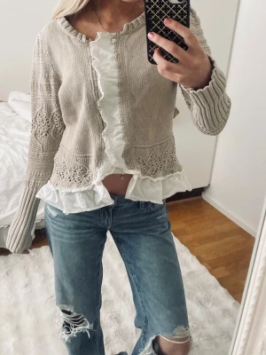 Beige kofta med spetsdetalj från Rosebud - Supersöt och unik beige kofta från Rosebud med  spetsdetaljer. Koftan har långa ärmar, ribbade muddar och vita volangkanter framtill och nedtill. Perfekt för lager på lager. Jag är 166 cm och när vanligtvis xs/s.