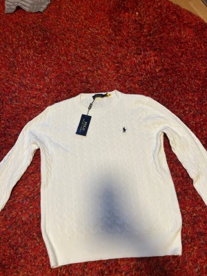 Vit kabelstickad tröja Polo Ralph Lauren - Kabelstickad vit tröja från Polo Ralph Lauren med klassisk rund hals och diskret logga på bröstet. Långärmad modell i mjuk bomull som passar perfekt till lager på lager. Enkel och stilren design som funkar till allt.