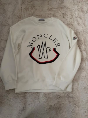 Vit sweatshirt från Moncler - Säljer en clean vit sweatshirt från Moncler med stort tryck på bröstet och logga på ärmen. Tröjan har rund hals, ribbade muddar och är tillverkad i mjuk bomull. Perfekt för dig som gillar stilrena och exklusiva plagg.