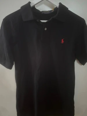 Svart pikétröja från Ralph Lauren - Klassisk svart pikétröja från Polo Ralph Lauren med röd broderad logga på bröstet. Modellen är custom slim fit och har korta ärmar, krage och knappar vid halsen. Perfekt till jeans eller shorts för en clean och stilren look.