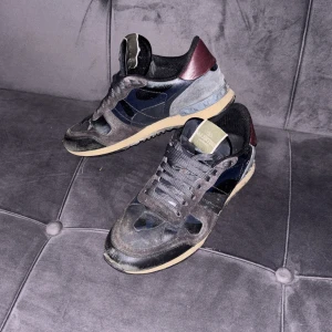 Valentino Rockrunner sneakers - Säljer ett par Valentino Rockrunner sneakers med cool camouflage-mönster i svart och mörkblå mocka och skinn. Skorna har vinröda detaljer vid hälen, beige sula och snörning framtill. Perfekt för dig som gillar lyxiga streetwear-vibes och snygga designer. Priset är ej hugget i sten och hör gärna av er vid frågor🤩🔥