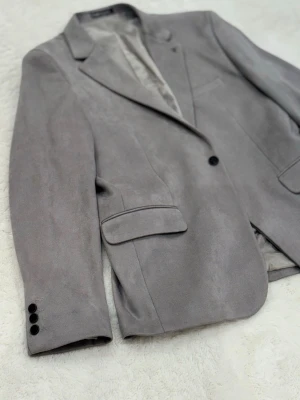 Grå mockakavaj från Mir Class - Snygg grå kavaj i mockaimitation från Mir Class. Klassisk modell med slag, tre knappar vid ärmslut och två fickor med lock framtill. Fodrad insida för extra komfort och stilren look. Perfekt för dig som vill ha en modern och clean vibe.