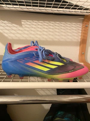 Adidas fotbollsskor i blått och rosa - Säljer ett par Adidas fotbollsskor med snygg färgkombination i blått, rosa och neongult. Skorna har snörning, låg profil och är tillverkade i syntetmaterial. Perfekta för dig som vill sticka ut på planen med stilrena och sportiga skor.