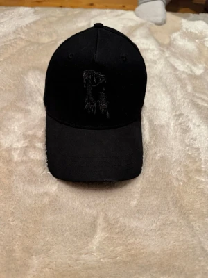 Svart keps från Risky med broderi - Säljer en svart keps från Risky med broderad logga framtill och texten 'RISKY' broderad baktill i en cool, droppande stil. Kepsen har böjd skärm och justerbart spänne bak. Perfekt för dig som gillar streetwear och vill ha en stilren accessoar.